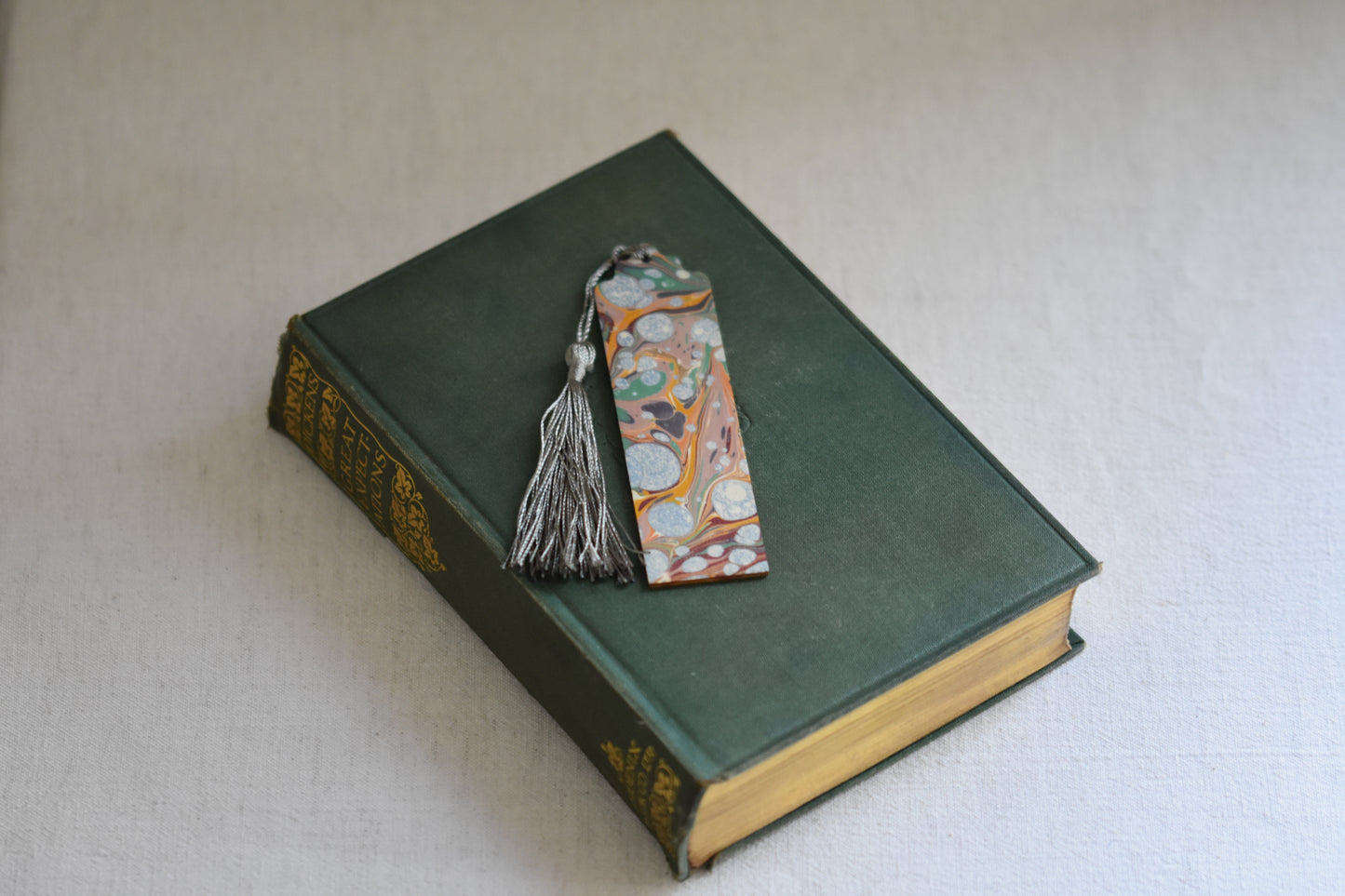 Jemma Lewis colourful marbled wooden bookmark with grey tassel on a vintage green and gold book, with a beige background.