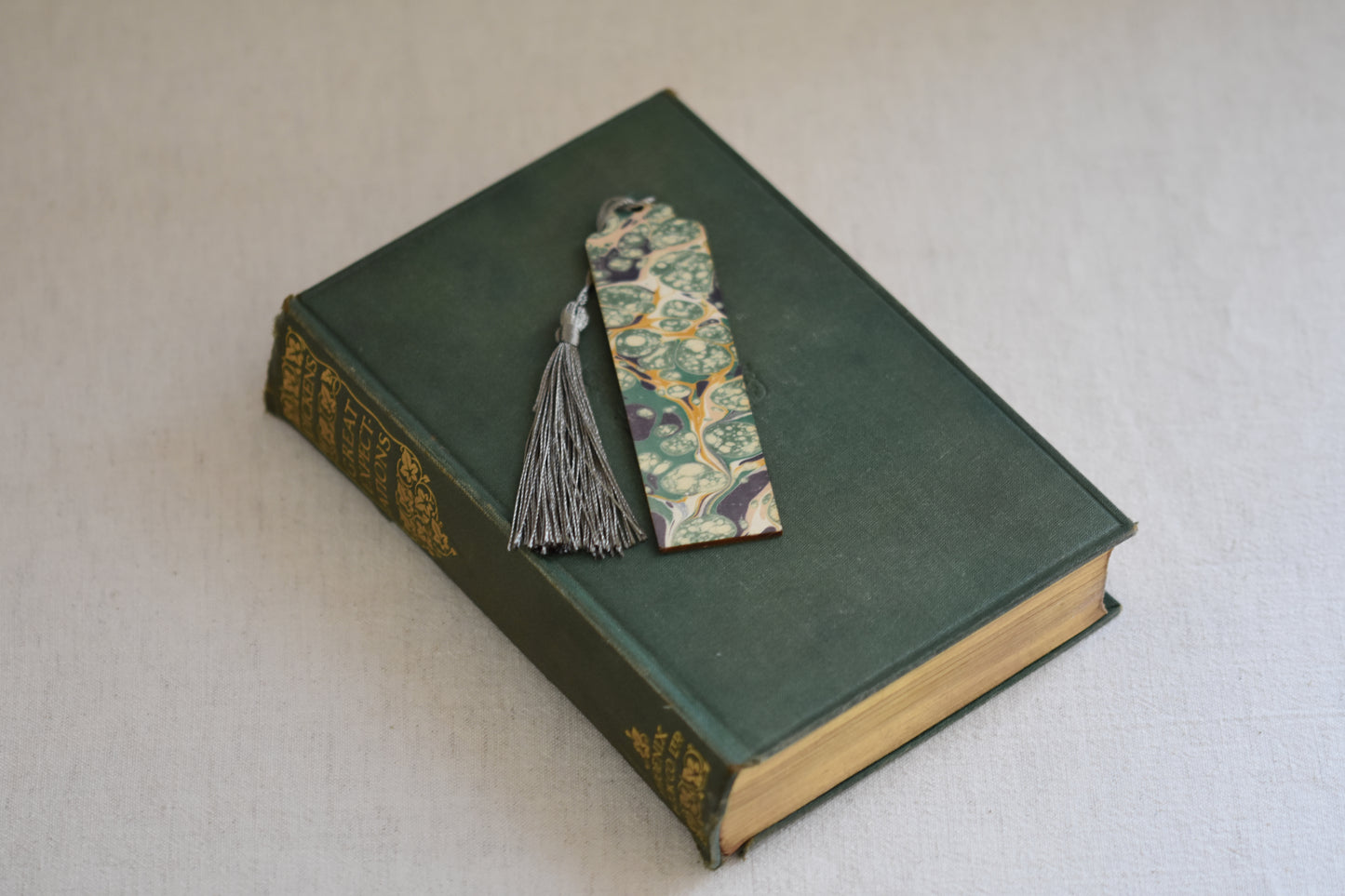 Jemma Lewis colourful marbled wooden bookmark with grey tassel on a vintage green and gold book, with a beige background.
