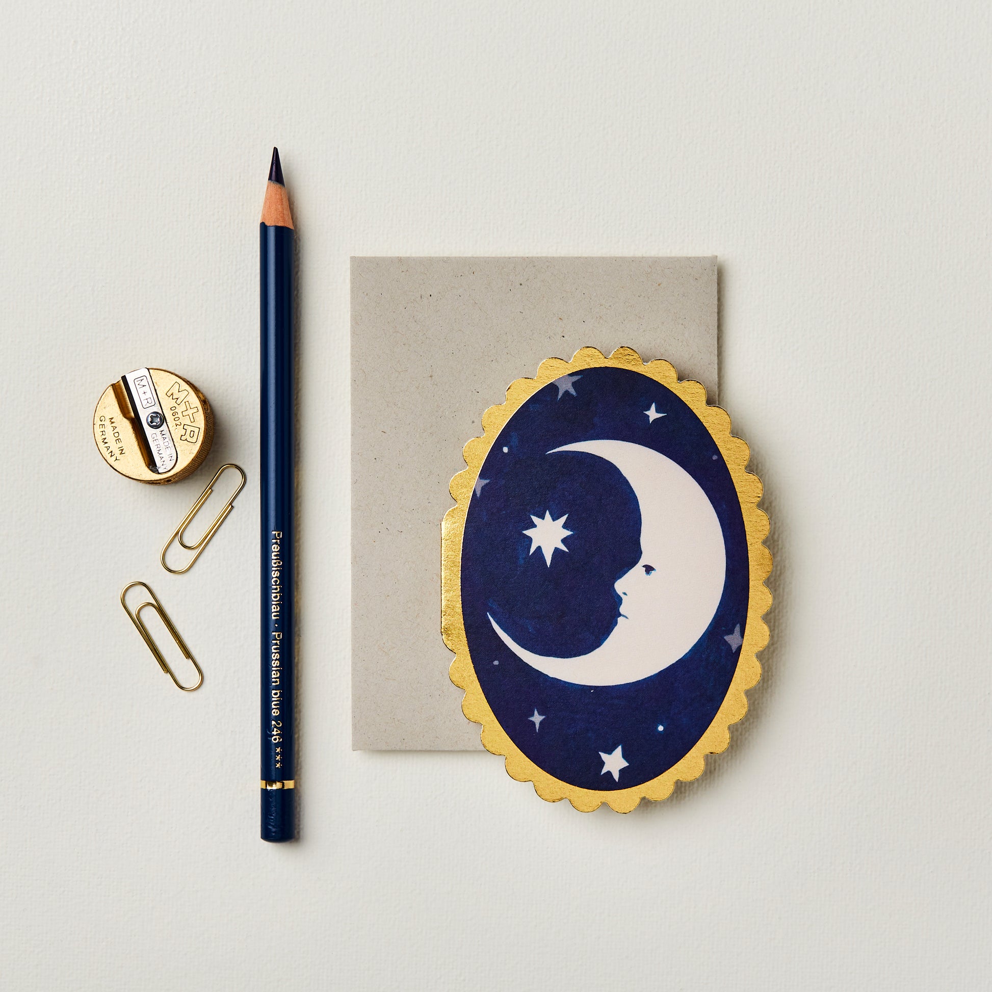 Wanderlust Paper Co. mini oval navy card with white moon design and gold scalloped edges, shown next to stationery on a pale grey background.