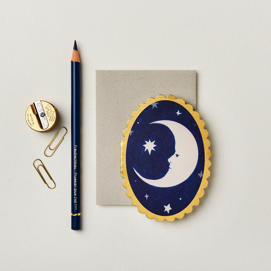 Wanderlust Paper Co. mini oval navy card with white moon design and gold scalloped edges, shown next to stationery on a pale grey background.