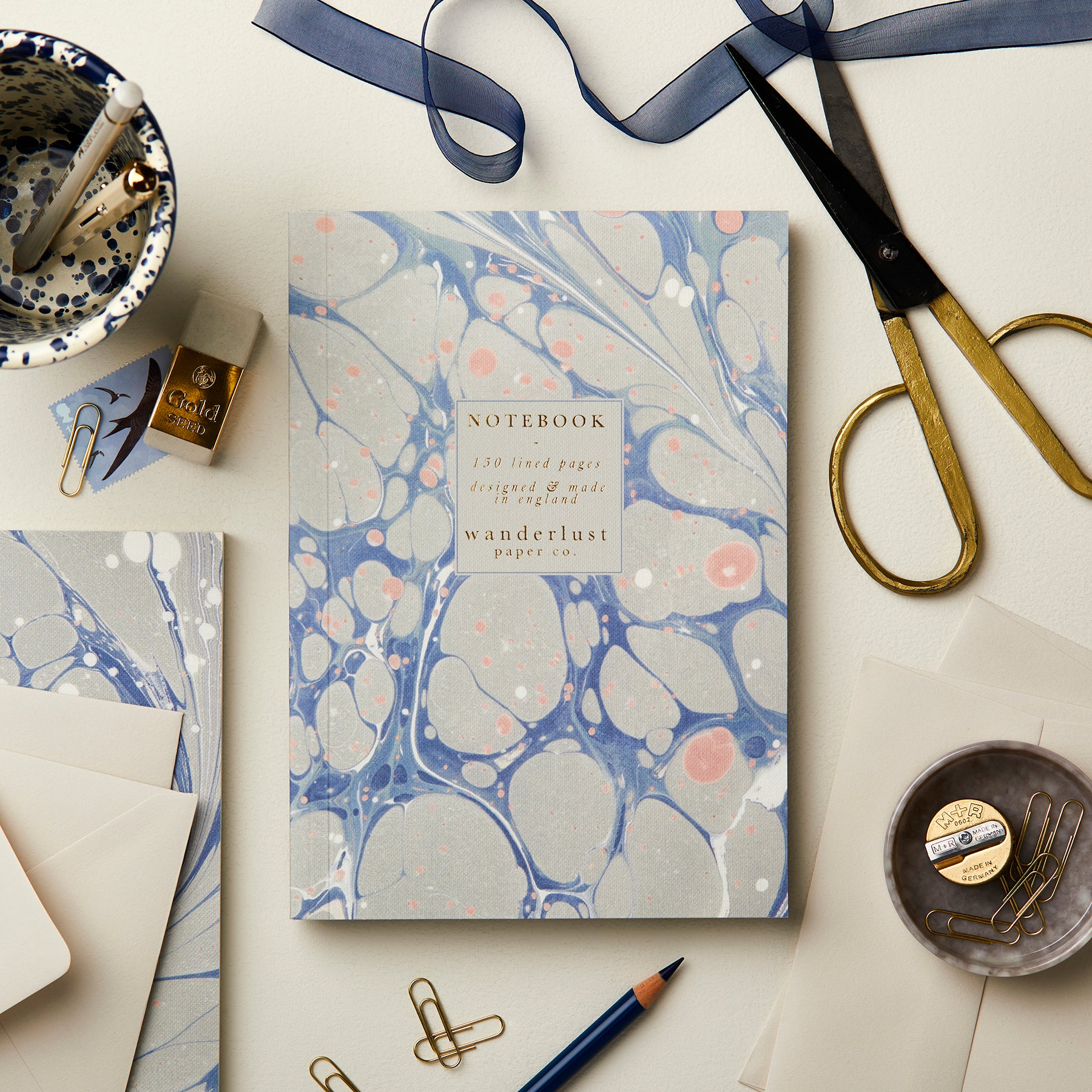 Wanderlust Paper Co. blue and pink marbled notebook, surrounded by stationery, on a light grey background.