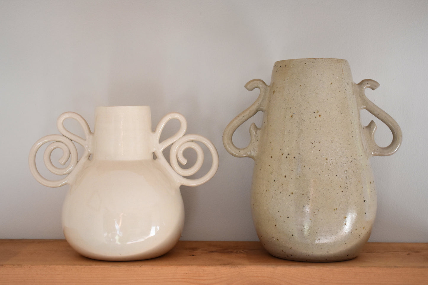 We Are Clay Pretzel Vase in white ceramic with spiral handles and Curly Vase in flecked beige ceramic with curly handles.