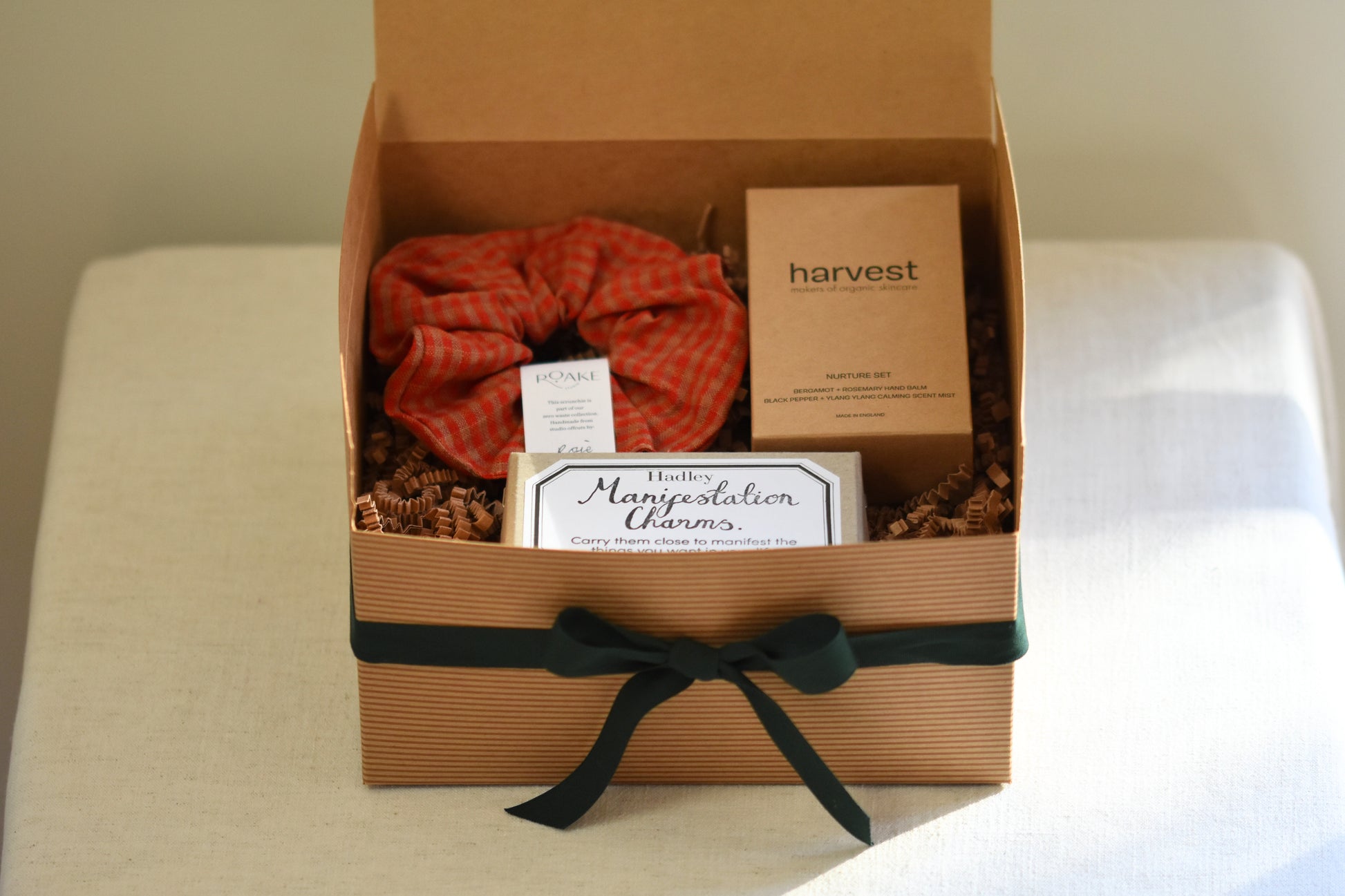RORA kraft gift box with green ribbon containing a Roake studio red scrunchie, Hadley manifestation charms and Harvest nurture set, on a beige background.
