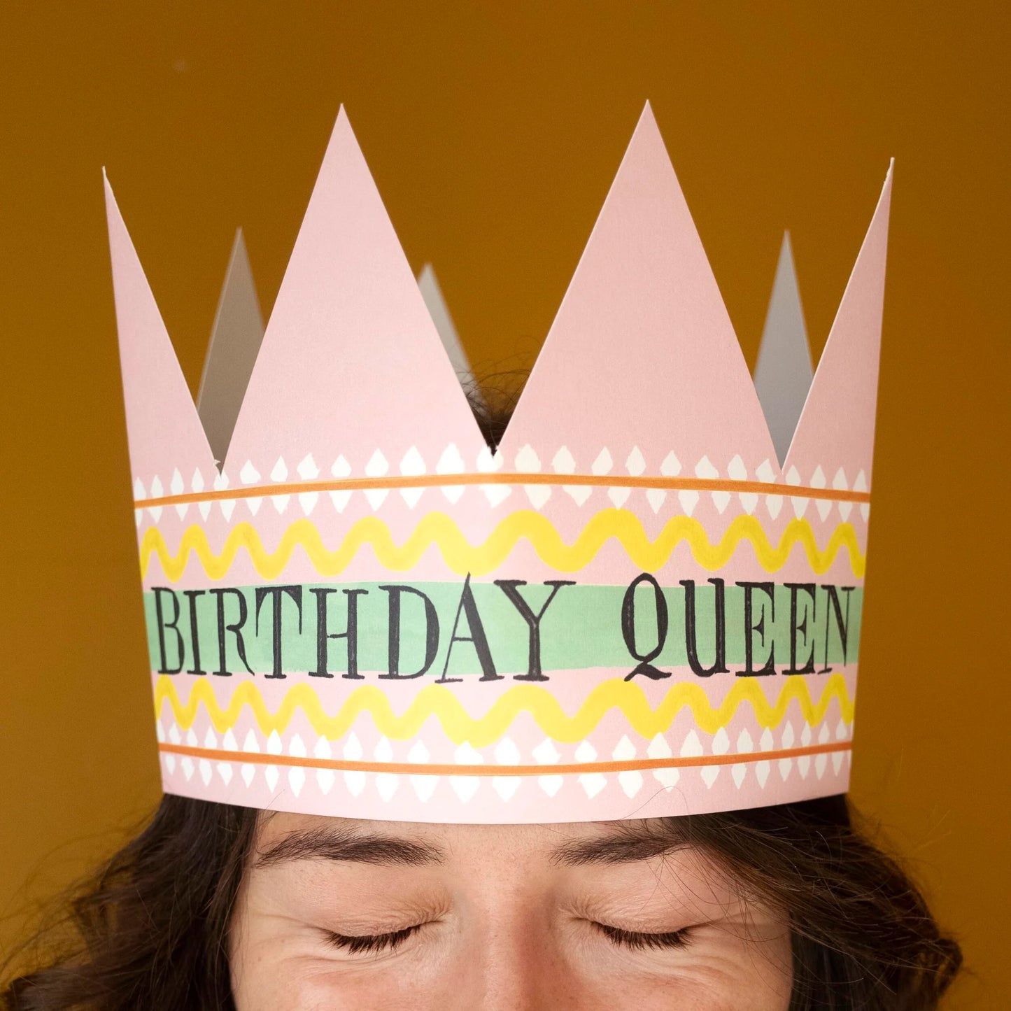 Hadley Paper Goods Birthday Queen party hat crown shaped card with pink, green and yellow pattern, worn by a person with a light brown background.