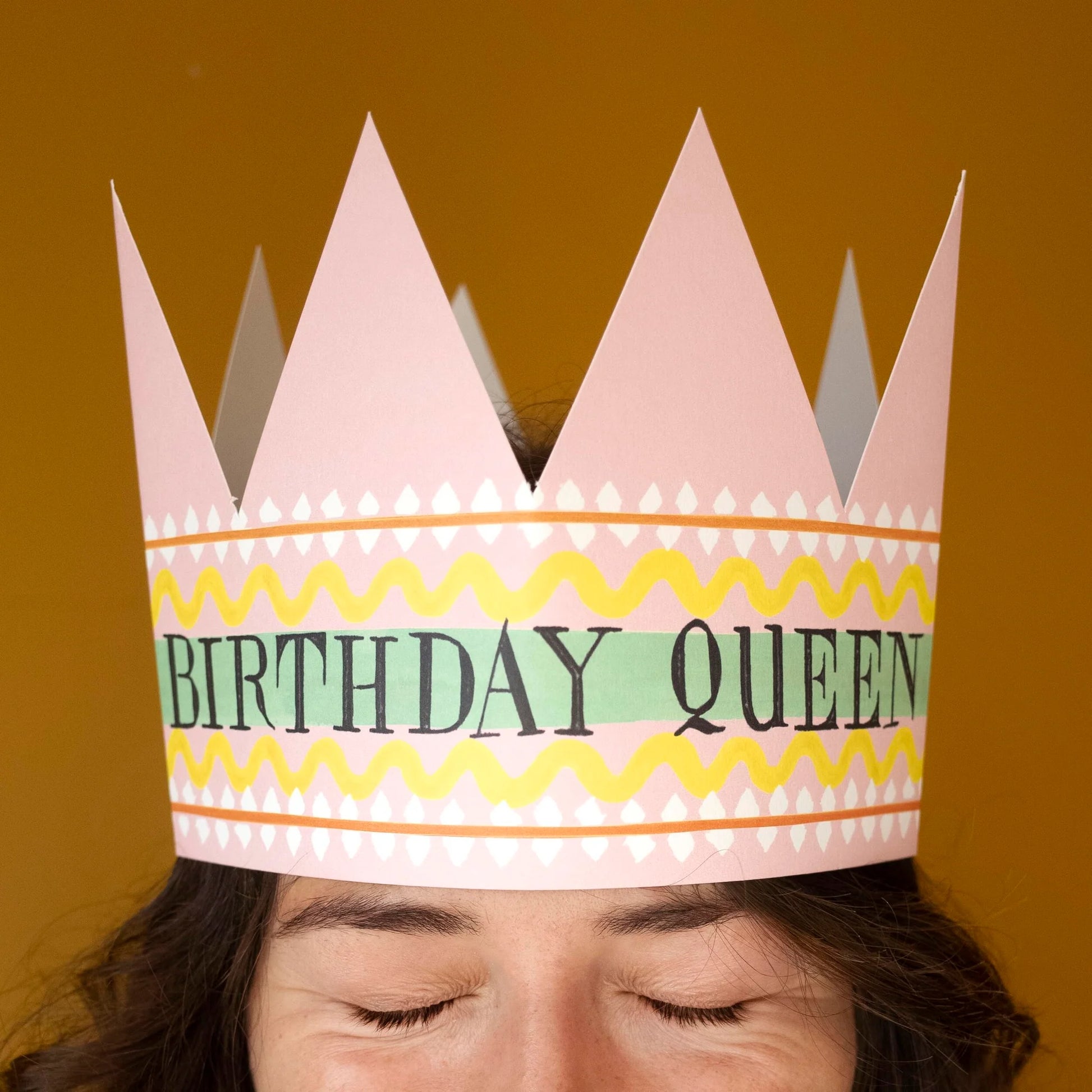 Hadley Paper Goods Birthday Queen party hat crown shaped card with pink, green and yellow pattern, worn by a person with a light brown background.