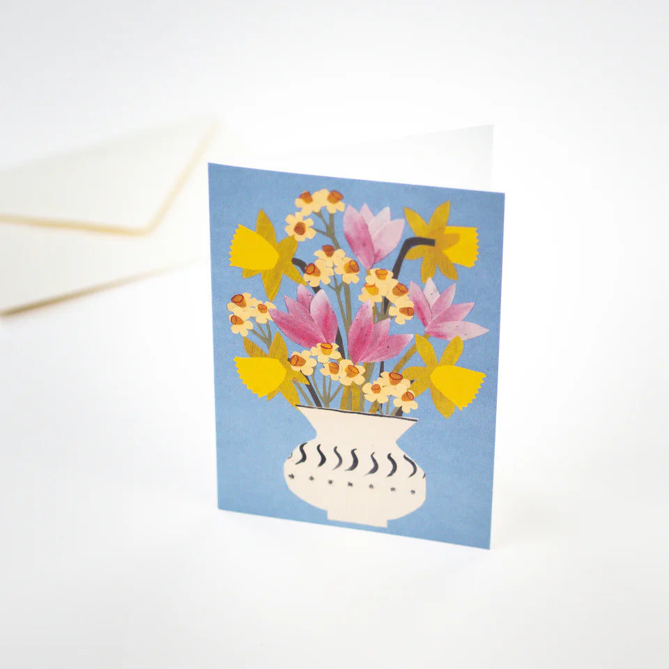 Hadley Paper Goods Spring flowers card showing a bouquet of daffodils, narcissus and tulips in a vase on a cornflower blue background with a cream envelope, on a white background.